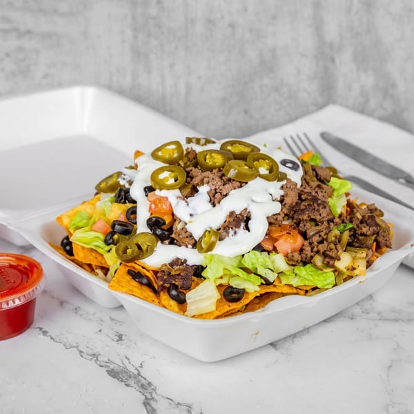 Loaded nachos with ground beef, lettuce, tomatoes, olives, jalapeños, and sour cream in a takeout container.