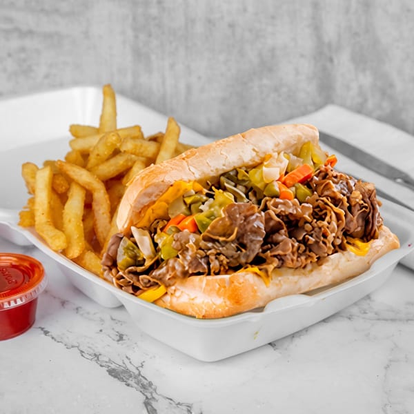 Cheesesteak sandwich with fries and two ketchup cups in a white takeout container on a marble surface.